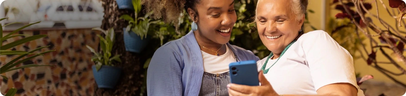 Duas mulheres sorrindo enquanto olham para um celular juntas em um ambiente com plantas.