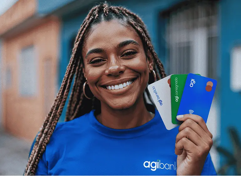 Mulher sorridente vestindo camiseta azul da empresa Agibank, segurando três cartões coloridos com a marca Agibank, ao ar livre em um ambiente urbano.